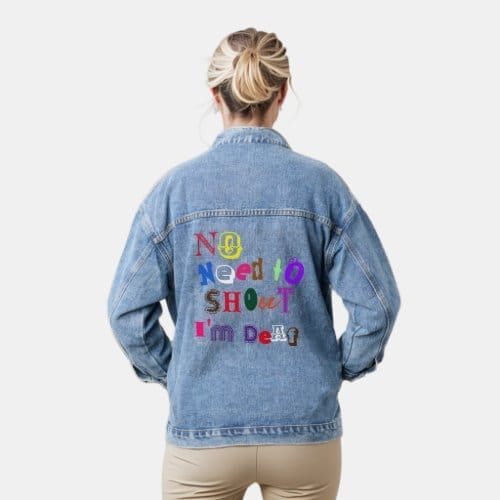 A woman wearing a denim jacket. On the back of the jacket in font from all different types, multicoloured, the words read, 'No need to shout, i'm deaf'