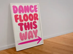 a print in a frame leaning against a wall. The print is all type, no picture the type is big and a bubble typeface, it is bright pink and reads, dance floor this way, with an arrow underneath